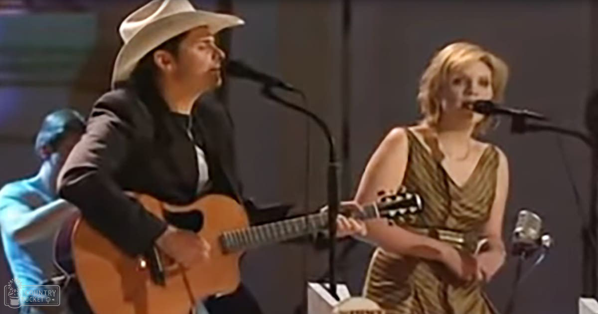 An adult male singer wearing a dark blazer, white shirt, black pants, and a light-colored cowboy hat performs on stage with an adult female in a shiny gold and brown patterned sleeveless dress. Microphones and guitars are present, suggesting a musical performance.