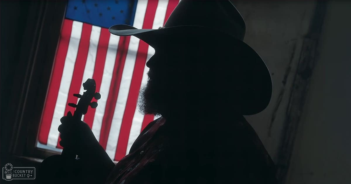 An older adult male singer wearing a wide-brimmed cowboy hat, dark-colored jacket with red accents, holding a violin against an American flag backdrop in what appears to be natural daylight.