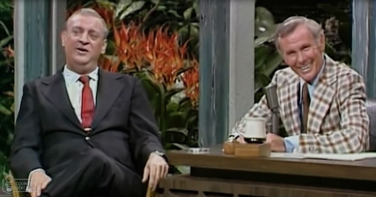 Middle-aged male talk show host wearing a dark suit and red tie interviewing another middle-aged man dressed in a plaid blazer over a light blue shirt, set against an indoor studio backdrop with large green plants and red flowers.