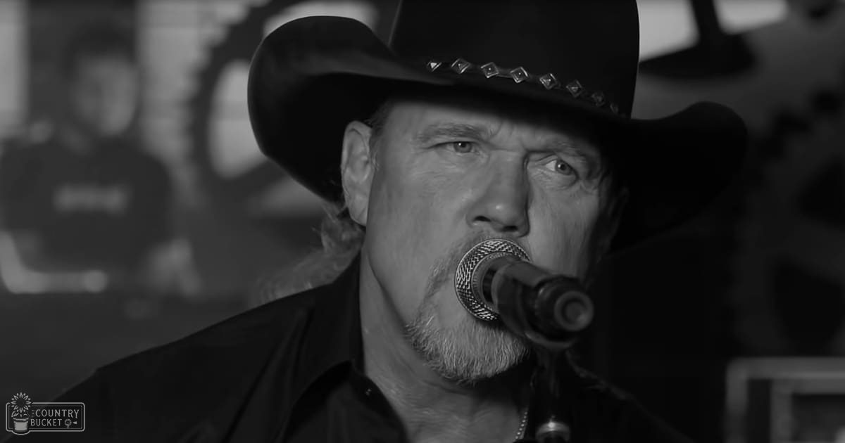 A middle-aged man wearing a black cowboy hat and dark shirt sings into a microphone on stage during an indoor performance.