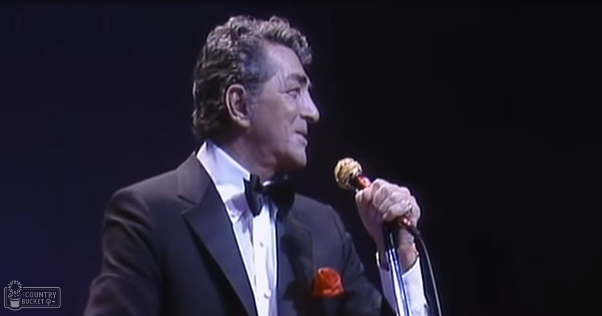 A middle-aged male performer in a dark tuxedo with an orange pocket square, singing into a microphone at what appears to be a concert venue.