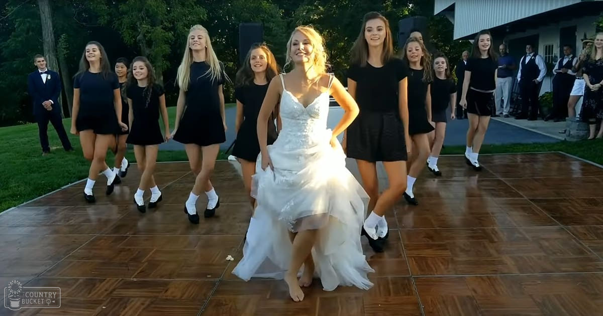 A bride in a white wedding gown dances with young women in black dresses at an outdoor wedding reception during late afternoon or early evening, surrounded by greenery and rustic elements.