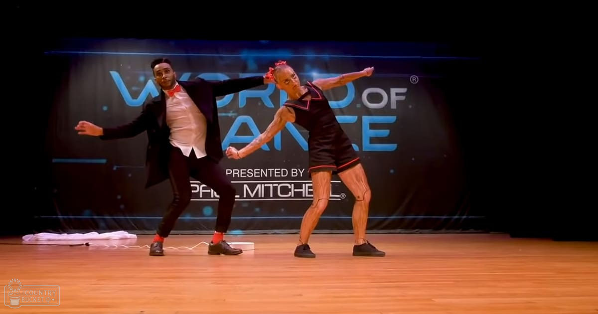 A dance performance at 'World of Dance,' presented by Paul Mitchell. Two young adults are on stage, one in a black suit jacket and white shirt with red socks, the other in athletic shorts and a tank top.