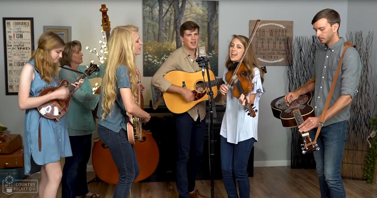 Group performing music indoors in a home studio or living room with various musical instruments visible.