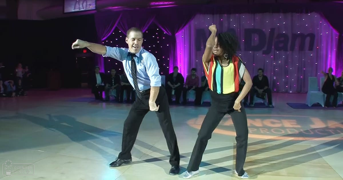 A dance performance at 'DJam' social event indoors with two adult dancers, a male and a female, performing on a checkered floor; stage curtains with purple lighting in the background.