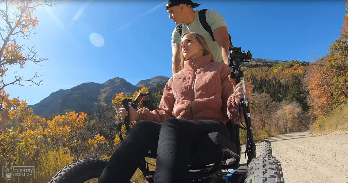 A young adult woman in a wheelchair with a man standing behind her on a dirt road amidst mountains, during autumn.