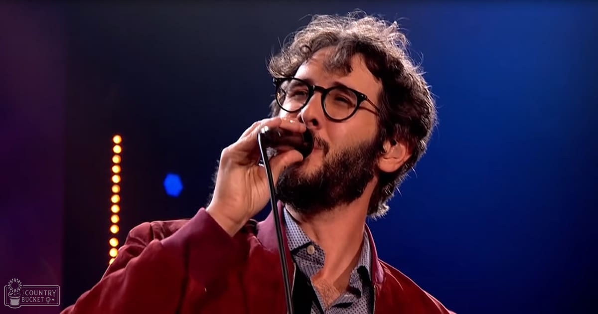 An adult male singer in a red jacket and patterned shirt sings into a microphone against a studio backdrop with vertical lights and blue lighting.