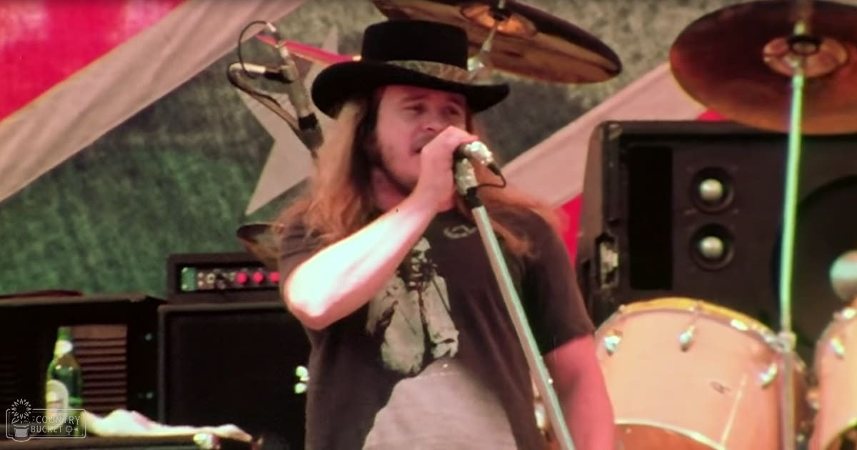 An adult male performer wearing a black cowboy hat and dark t-shirt with a graphic print, singing into a microphone at an outdoor music festival.