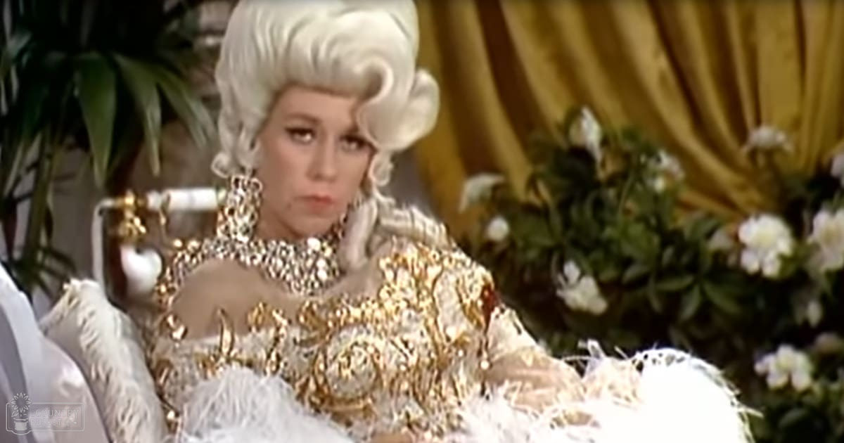 An adult female wearing an ornate gown with intricate gold embroidery and feather embellishments stands in a luxurious indoor setting with greenery and golden accents.