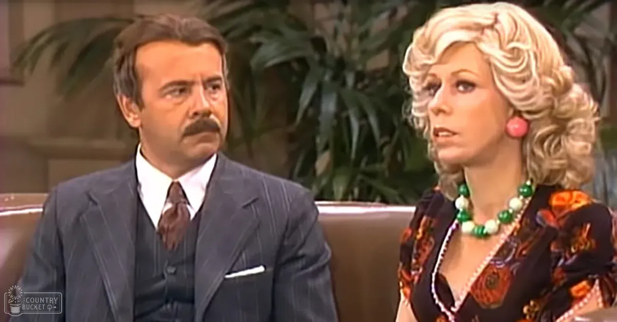 Indoor setting of a mid-20th century television production with two middle-aged individuals in formal attire and casual fashion, surrounded by indoor plants under soft studio lighting.