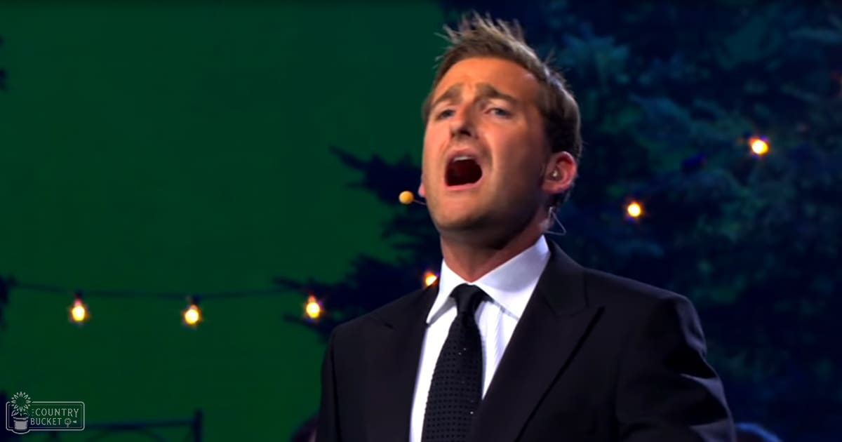 A young adult male singer wearing a black suit jacket and white dress shirt performs on an indoor stage with green screen and string lighting, suggesting a live performance or event.