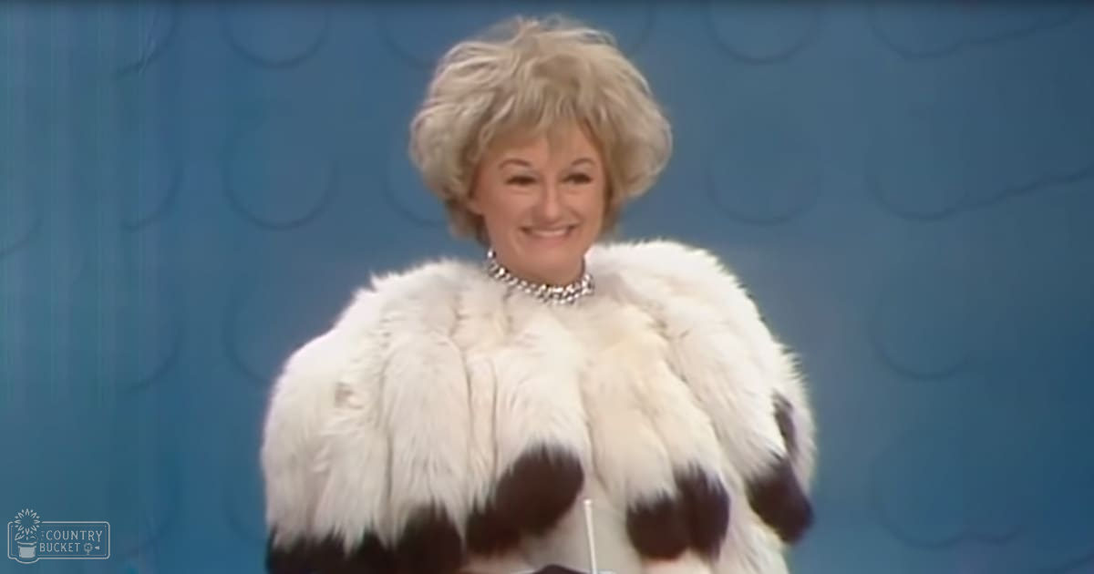 Older adult female in a studio setting, wearing a white fur coat with black accents and a silver necklace against a blue abstract background.