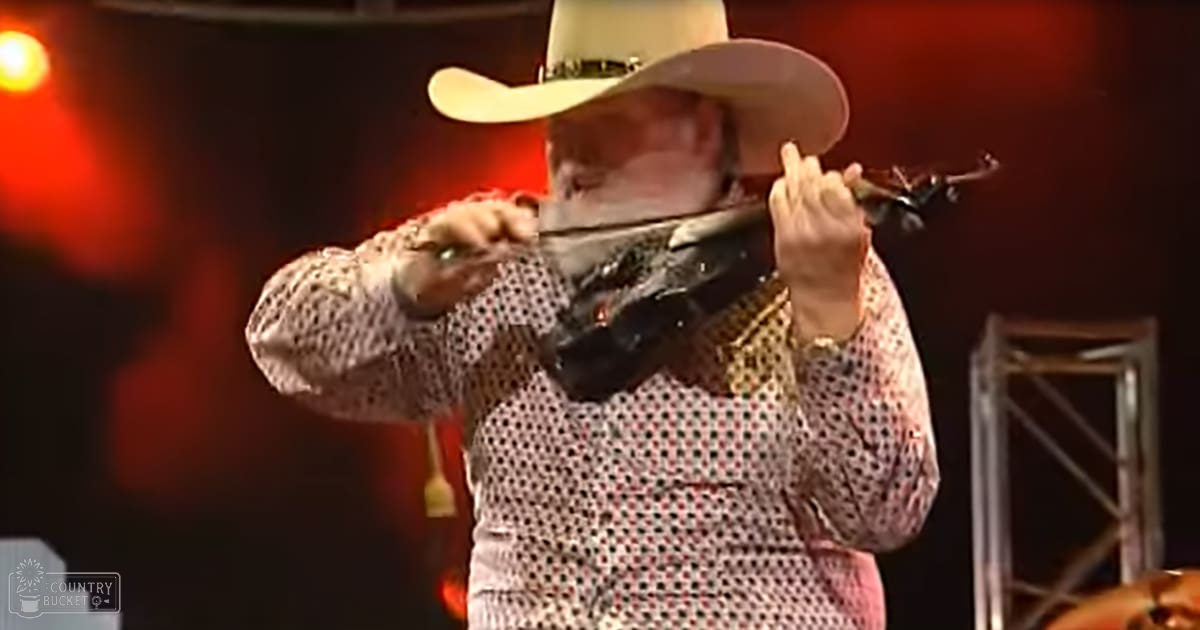A middle-aged male musician wearing a white cowboy hat, polka dot shirt, and gold jewelry performs with a violin on stage under red and orange lighting.