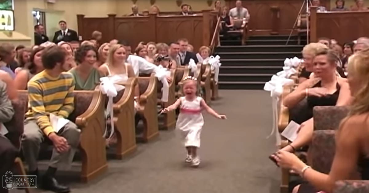 A young child running down an aisle in a church setting, surrounded by seated adults dressed formally for what appears to be a wedding or ceremony; the scene is lit with natural light.