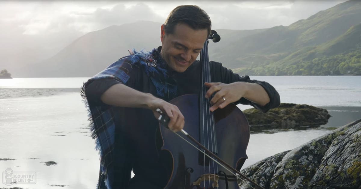An adult male playing a cello outdoors near water with mountains in the background, wearing a dark plaid jacket under bright daylight conditions.