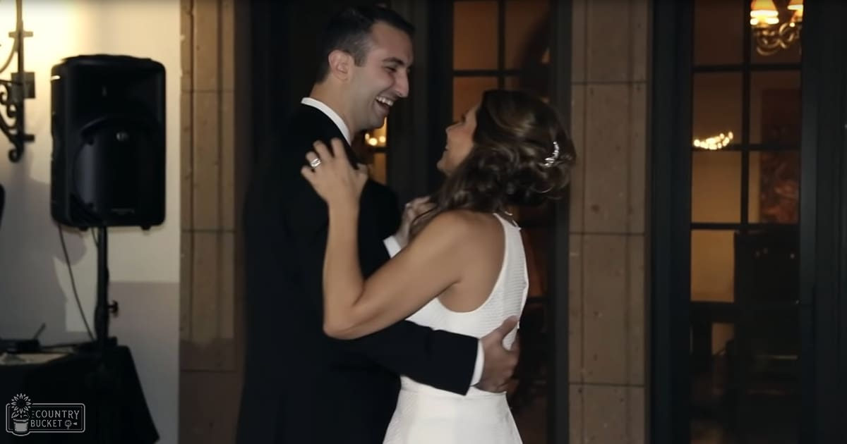 A couple dancing indoors at an event or reception, wearing formal attire; male in a black tuxedo with white dress shirt, female in a white sleeveless patterned dress, both smiling and holding each other closely under artificial evening lighting.