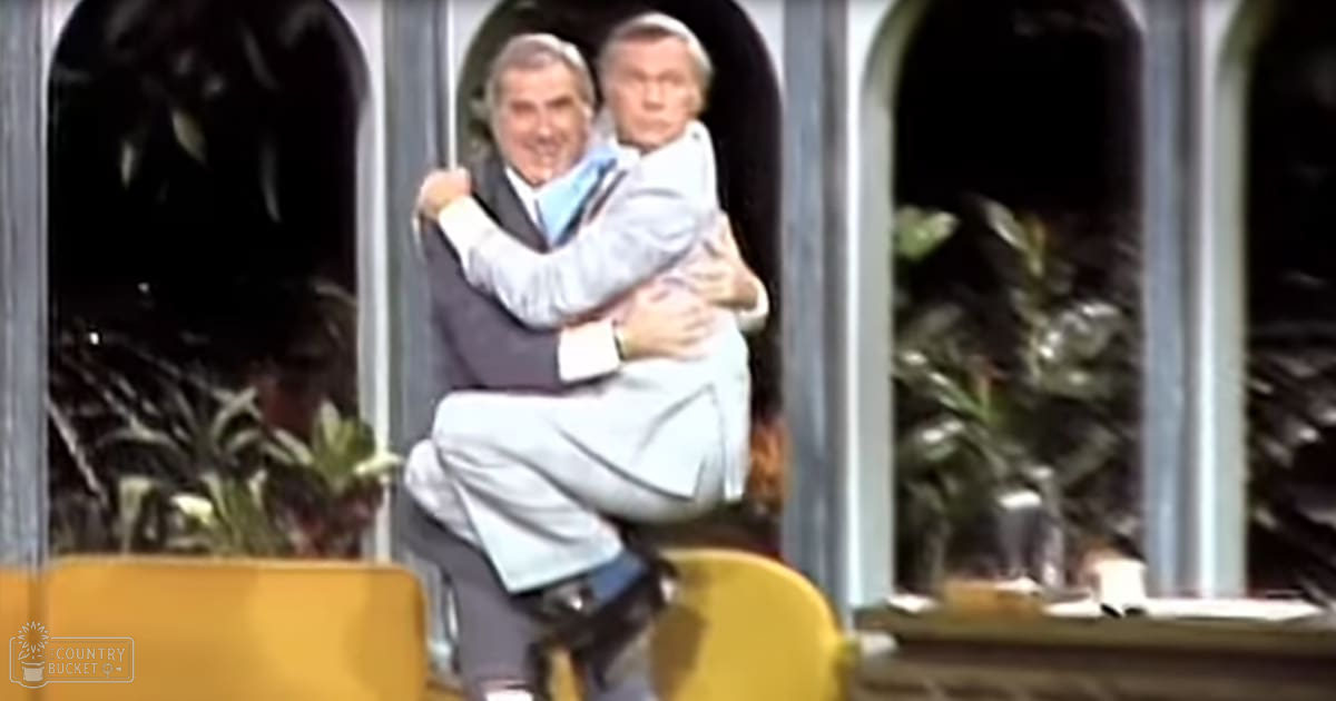 Two older adult males in light-colored suits sitting indoors with yellow chairs and greenery visible through arched windows, suggesting a spring or summer daytime setting.