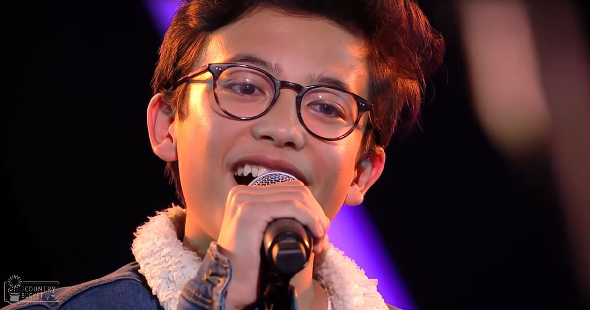 A young adult male in a denim jacket with a fluffy white collar sings into a microphone on stage under dim, purple-lit conditions.