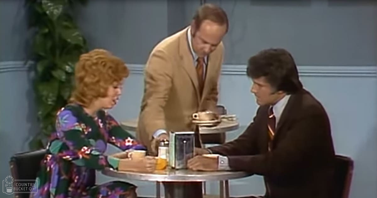 A studio talk show set with an adult female in a floral dress, an adult male in a beige suit jacket, and another adult male in a dark brown suit jacket. The backdrop includes a light blue wall and a decorative plant.