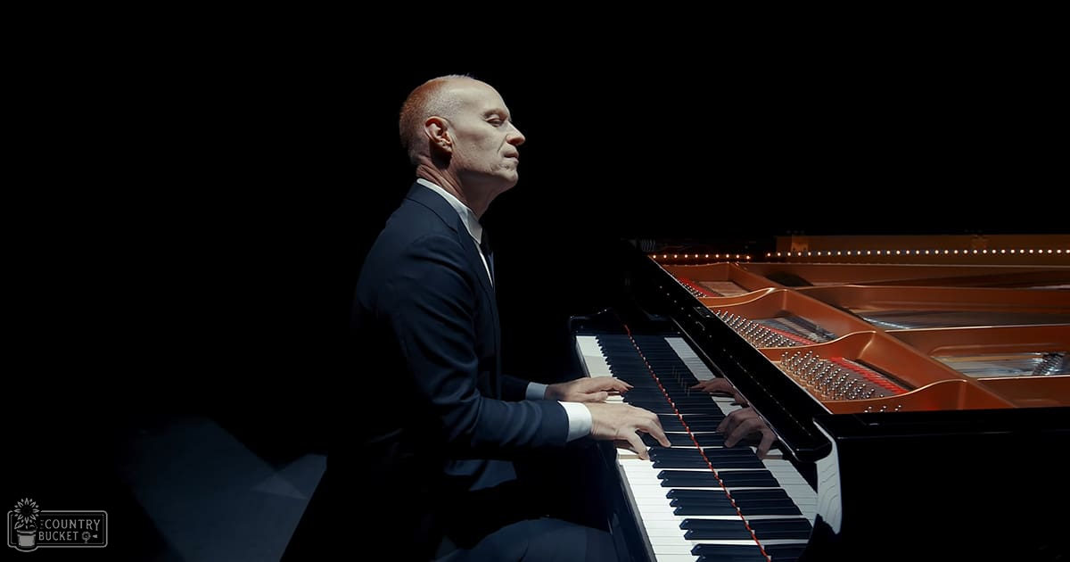 A middle-aged adult male plays a grand piano in an indoor concert hall setting with dim stage lighting.