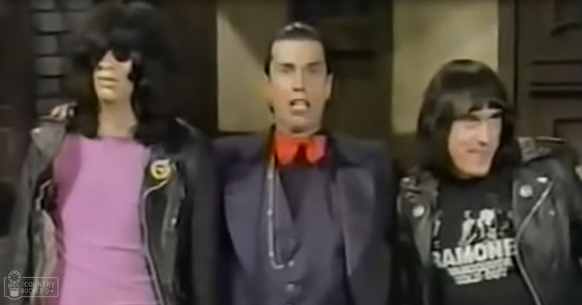 Adult female wearing a light pink dress with a fitted cut and high neckline, paired with a black leather jacket over it; adult male in a dark suit with a red bow tie and white shirt underneath; young adult female in a black t-shirt reading "RAMONE" with graphic designs, also wearing a black leather jacket. Indoor setting with even lighting.