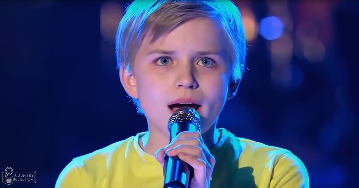A young adult child sings into a microphone on an indoor stage with dark stage lighting.