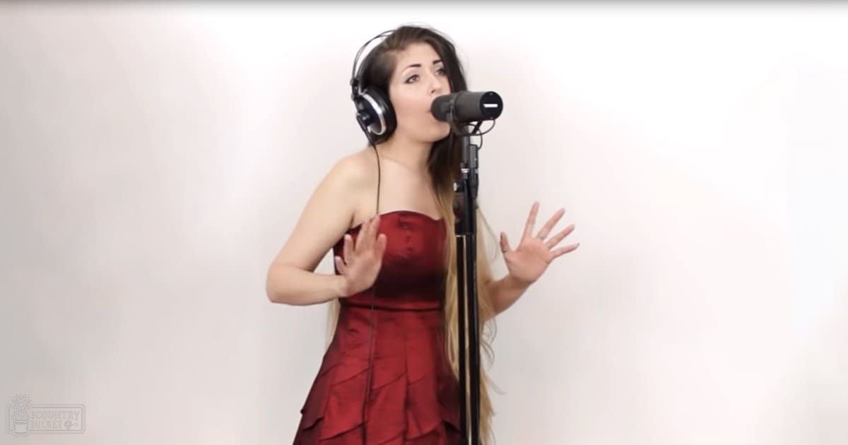 A young adult female wearing a strapless red dress with ruffled details, form-fitting and elegant, indoors while holding large over-ear headphones. Microphone on a stand suggests a recording or broadcasting setup against a plain white background.