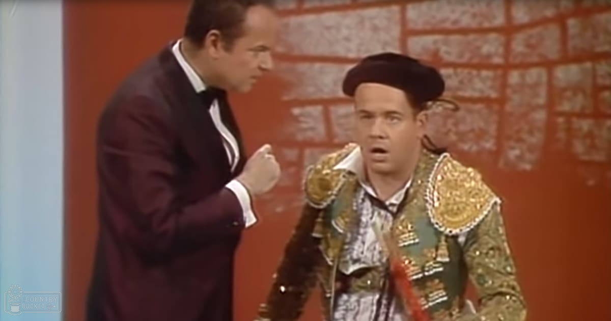 A man in traditional bullfighter attire stands next to another person, both indoors with a brick-like background. The scene appears well-lit during daytime or by studio lights.