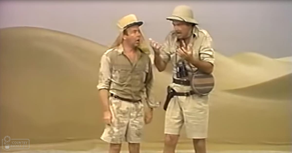 Two men in a desert setting wearing beige or light brown shirts and shorts, wearing safari-style hats under bright daylight.