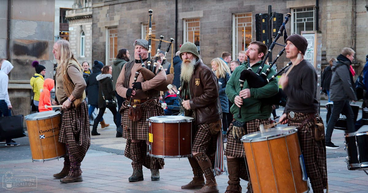 A street performance with bagpipers and drummers in traditional Scottish attire against historic urban architecture.