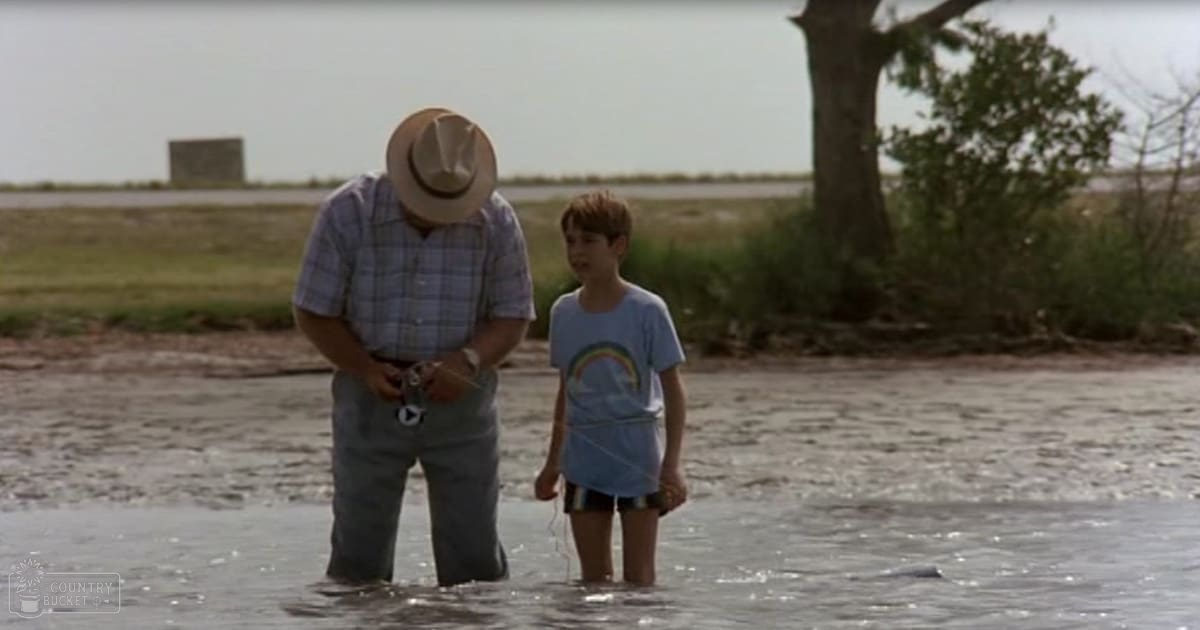 Two people near water holding fishing rods, wearing casual clothing and a straw hat under overcast skies.