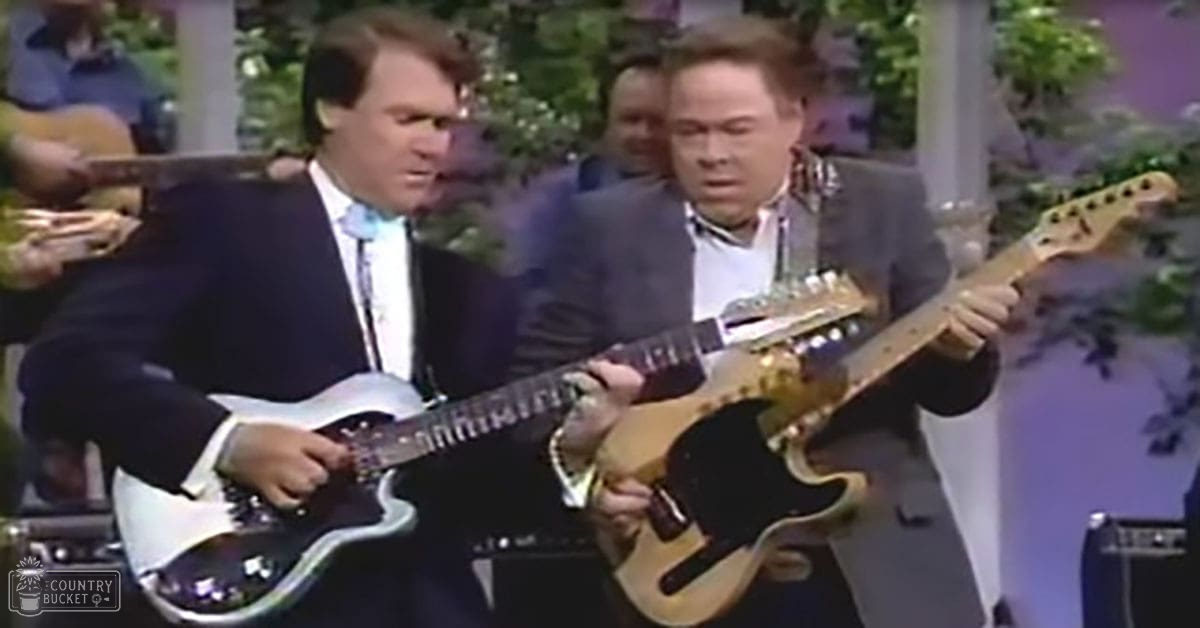 Indoor stage setting with two adult male musicians performing, surrounded by greenery and plants.