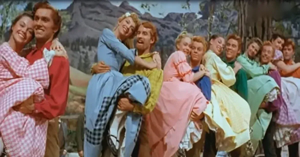 A group of young adults performing on a stage with painted mountain and tree backdrop, bright daylight lighting.