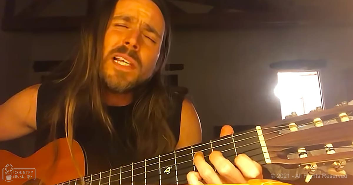 An adult male playing a guitar indoors, wearing a sleeveless black top with natural light streaming in from a window. The setting appears to be a home or studio environment.