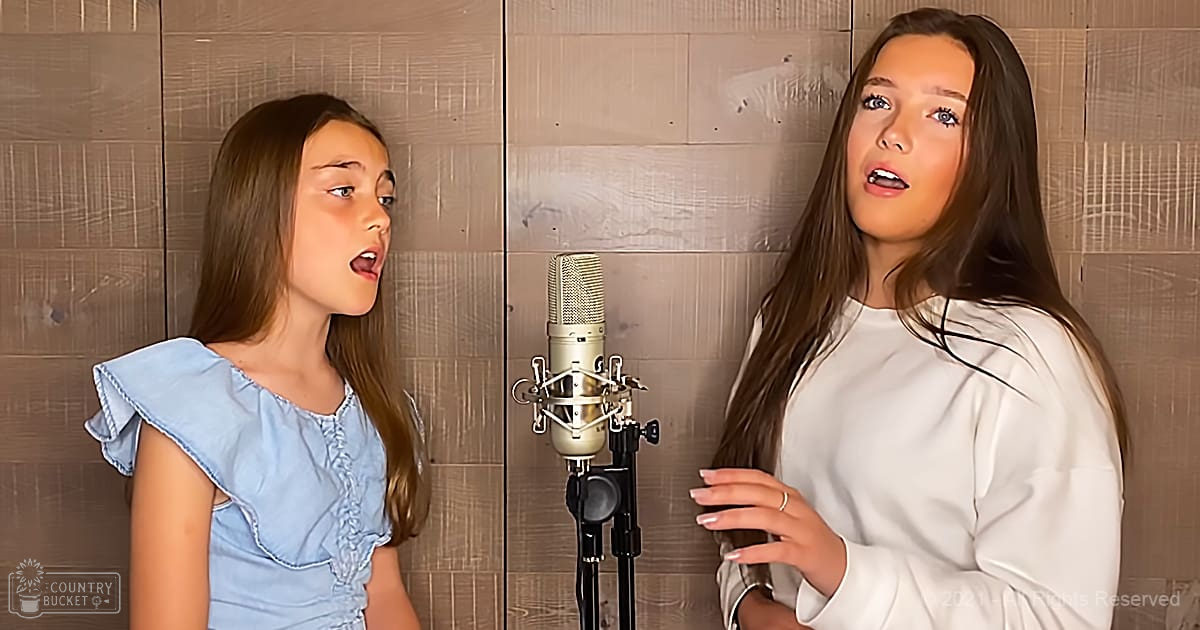 Two individuals in a recording studio setting, one child/teen girl wearing a light blue ruffled denim-like dress, another young adult woman in a white long-sleeved silk or satin top. Wooden paneling and professional microphone visible.