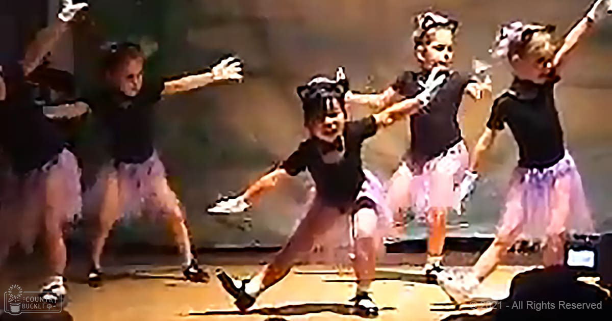 Children performing a dance routine in an indoor stage setup with simple backdrop and lighting equipment, wearing black sleeveless tops and light purple tutu skirts.