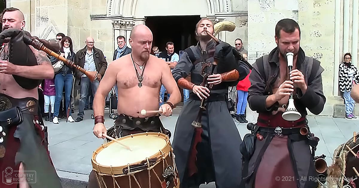 A group of adults performing traditional music on bagpipes and a drum in front of an old stone building, surrounded by spectators dressed for cooler weather.