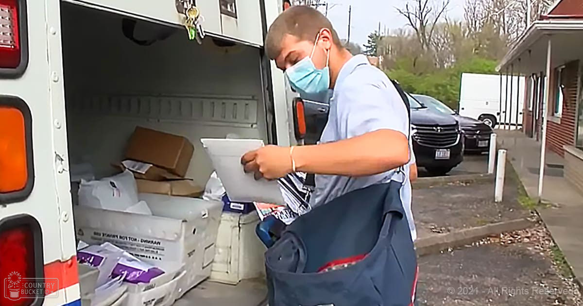 Young adult male wearing a light blue graphic t-shirt with relaxed fit, crew neck, short sleeves, face mask, watch or bracelet, standing near an urban delivery van filled with packages and boxes.