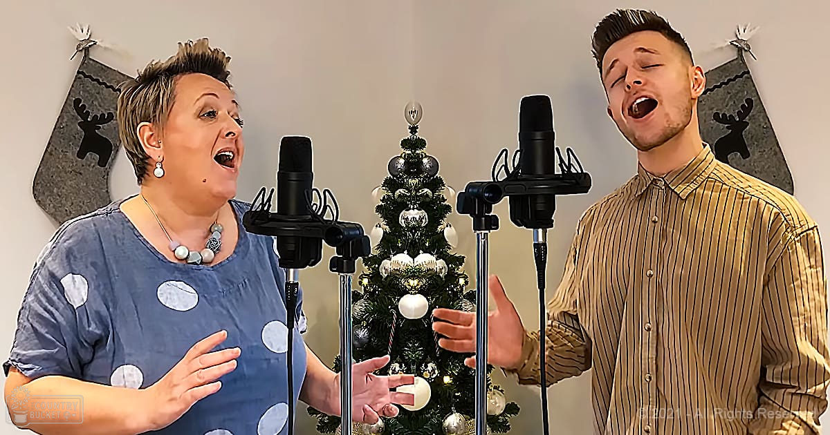 Adult female in a blue polka-dot blouse with large earrings and beaded necklace duets with young adult male wearing striped button-up shirt indoors during the holiday season, in front of a decorated Christmas tree.