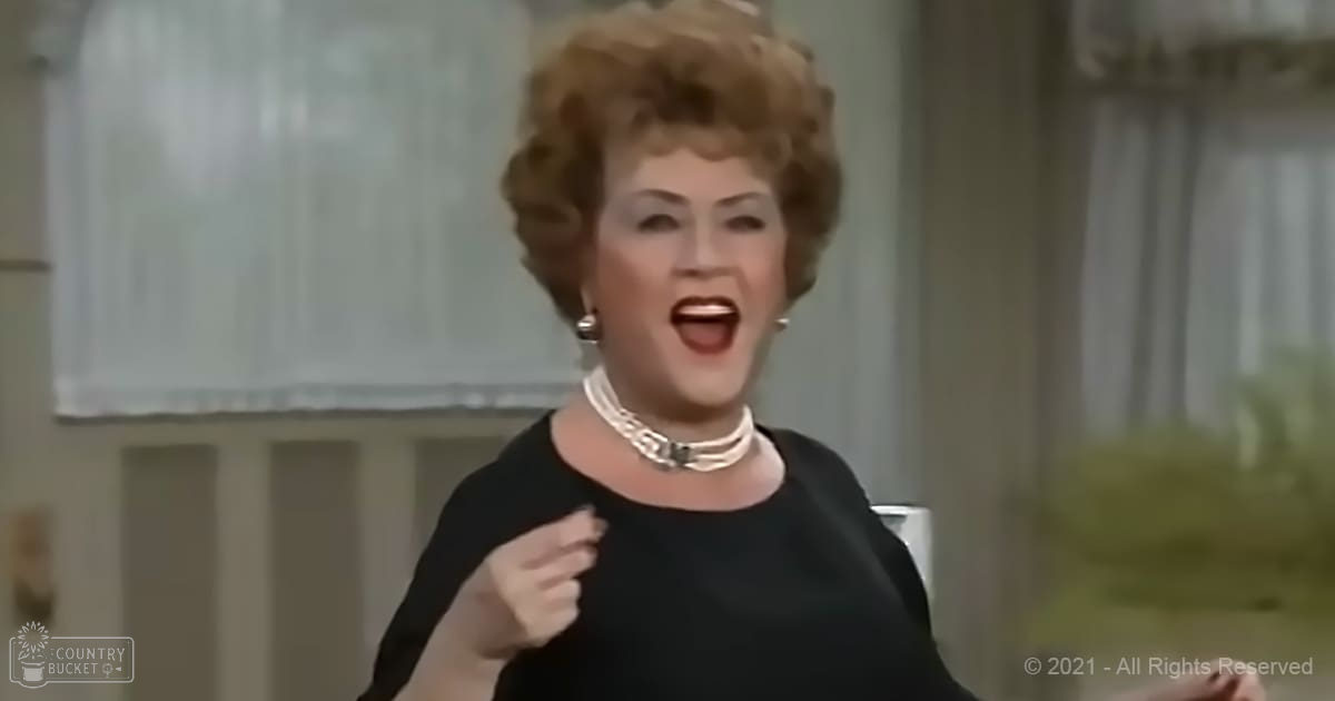 A middle-aged adult female in a form-fitting, solid black dress with short sleeves, accessorized with a choker-style necklace and earrings, stands indoors by curtains and a window.