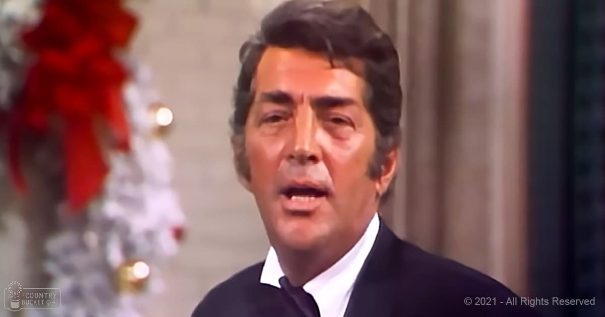 Middle-aged adult male in a dark suit and white shirt, standing indoors with a blurred Christmas tree decorated with red ribbons and white ornaments in the background.