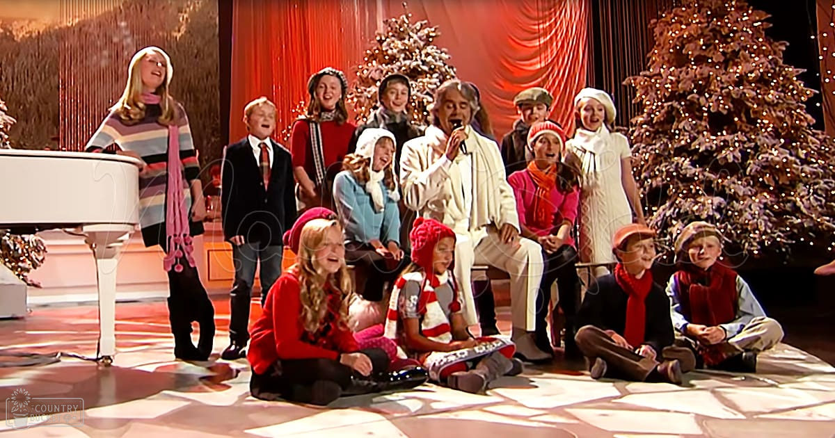 Group performance during Christmas time with adults and children in festive attire, white grand piano on the left, Christmas trees in the background, patterned carpet or floor.
