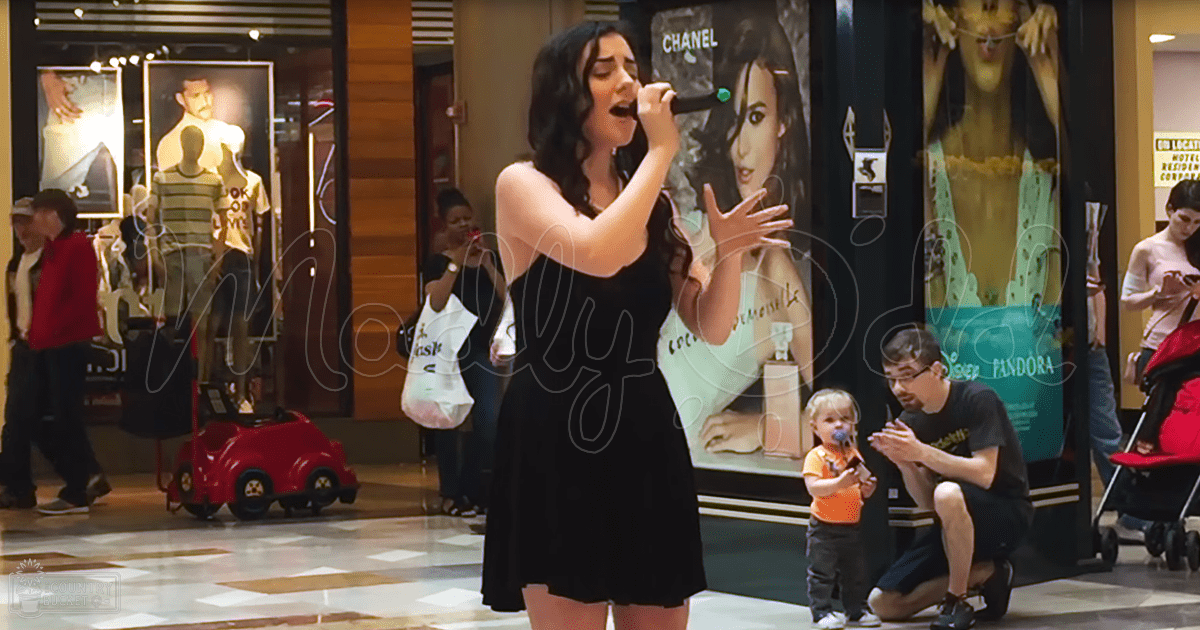 A young adult female singer wearing a black sleeveless dress performs in what appears to be a shopping mall. A toddler and an adult male are also present, both casually dressed.