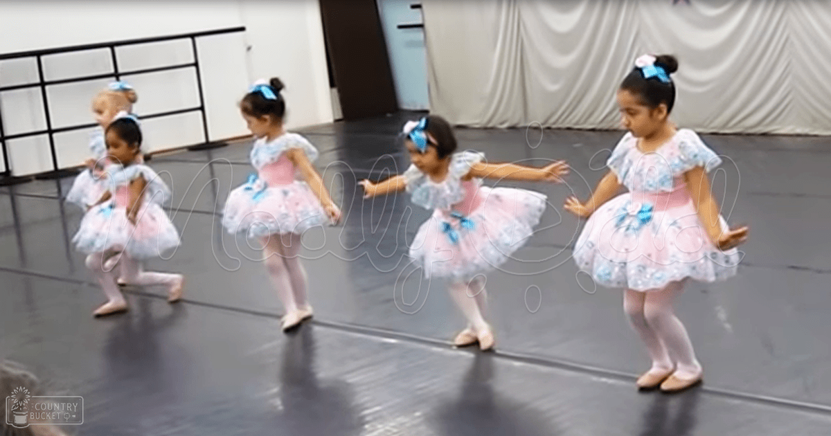 A group of young children, primarily female toddlers to early school-age girls, dressed in ballet costumes with tutus, tights, and ballet shoes, are practicing or performing in a dance studio with black flooring, railing, and white drapes.