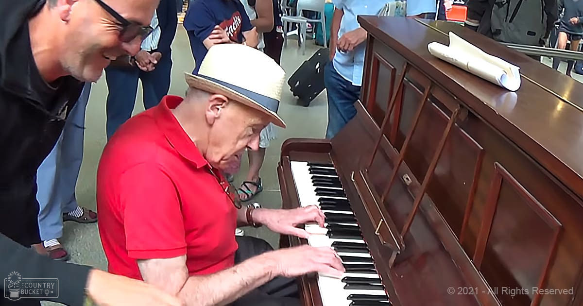 An older adult male in a red polo shirt plays a brown wooden upright piano in an indoor public space with plastic chairs and natural daylight.