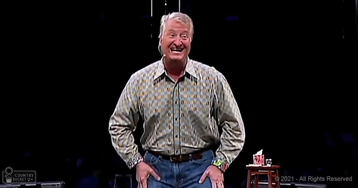 An older adult male in a performance setting, wearing a long-sleeved button-up shirt with muted beige and green patterns on a light background, standing near a dark stool with a drink atop it.