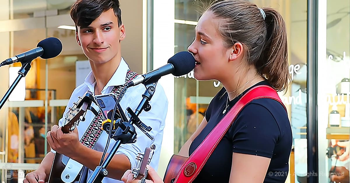 A young adult male and female duo performing music with microphones on stands in an indoor shopping mall setting.