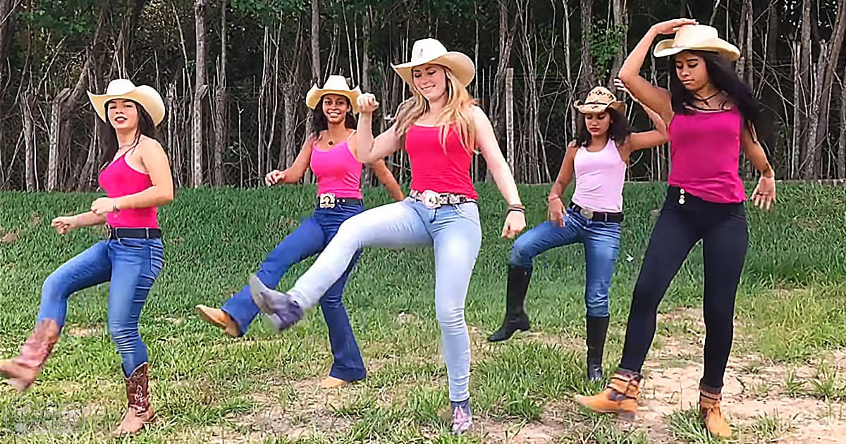 Five young adult women dancing outdoors in a rural setting with grassy terrain and trees, wearing pink tank tops and jeans or leggings, under soft afternoon light.