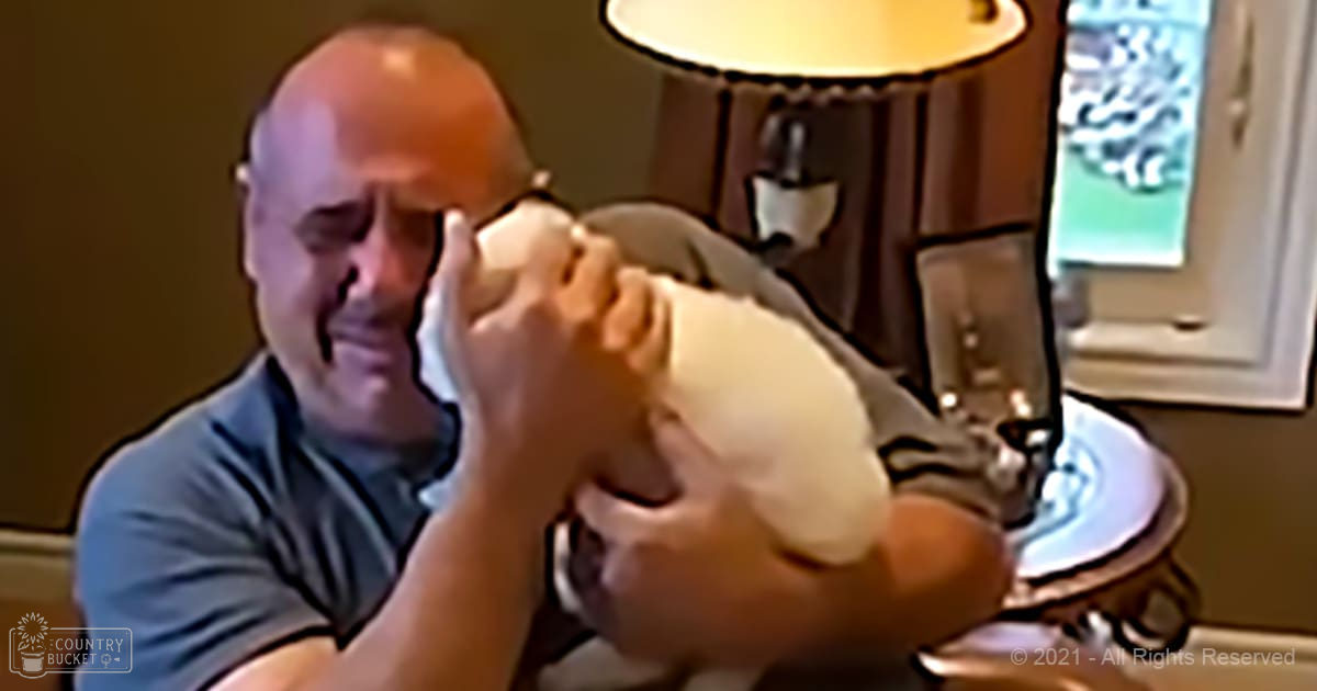 A middle-aged man holds an infant in a home setting with beige walls, daylight streaming through the window. The man wears a gray crew-neck t-shirt with black stripes on sleeves and collar.