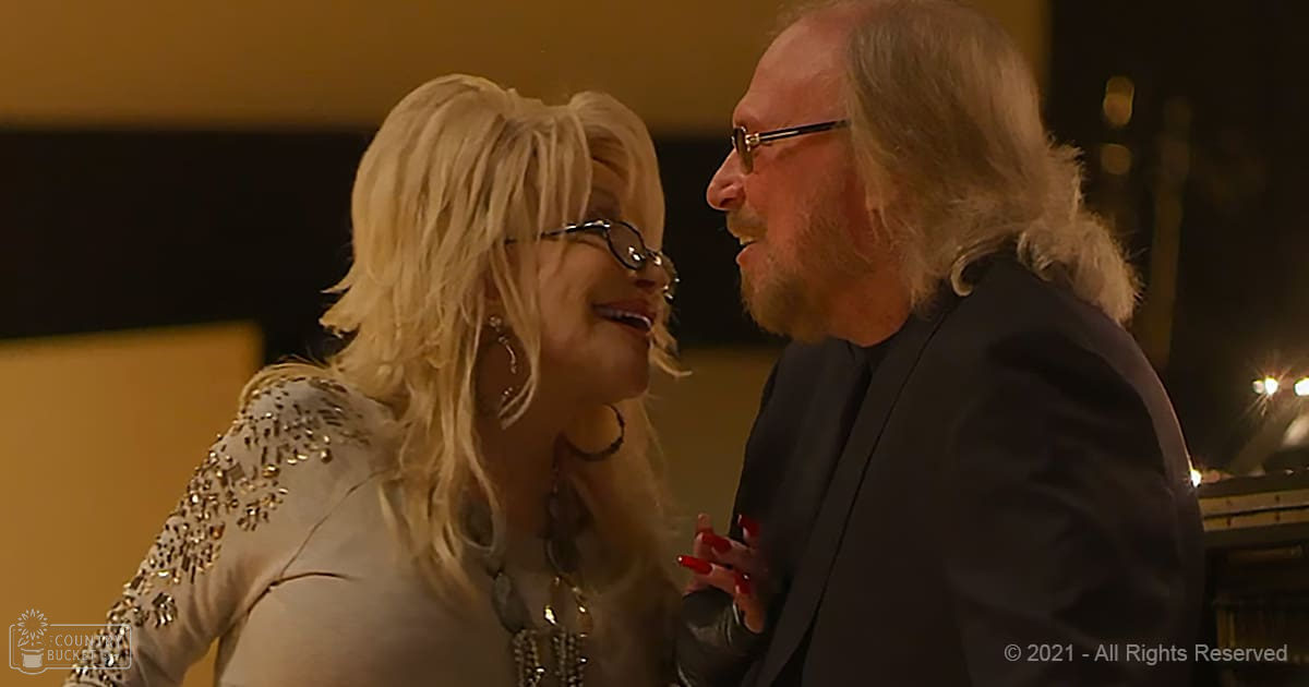 A woman wearing a sequined white top and layered necklaces, accompanied by a man in a dark suit jacket, with warm indoor lighting suggesting an evening setting.